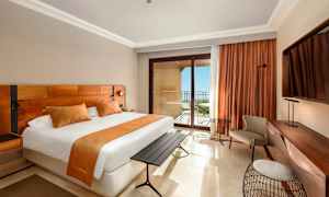 Et hotelværelse med en stor seng, orange indretningsdetaljer, et skrivebord og en altan med havudsigt. (AI-genereret tekst)