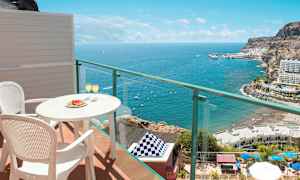 Udsigt fra altan med bord og to stole, hvor glas er serveret, mod havet og kystlandskab. (AI-genereret tekst)