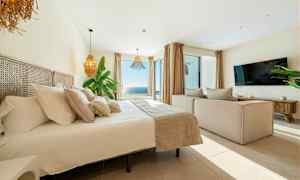 Billede af et lyst og moderne soveværelse med stor seng, sofa, TV og en bred udsigt over havet gennem store vinduer. (AI-genereret tekst)