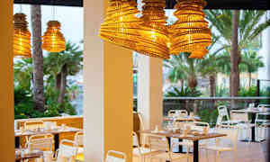 Udendørsmøbler ved restaurant med hængende lampeskærme af træ og palmer i baggrunden. (AI-genereret tekst)