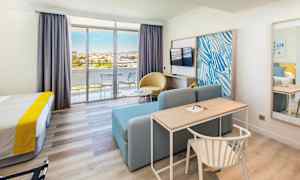 Et hotelværelse med seng, skrivebord, sofa, tv og glasdøre, der fører ud til en altan. (AI-genereret tekst)