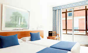 Et hotelværelse med to enkeltsenge, der har blå tæpper og puder, en stol ved et bord samt et vindue med blå gardiner. (AI-genereret tekst)