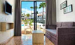 Udsigt over en stue med sofa og bord samt en åbning til en balkon med udsigt over en pool. (AI-genereret tekst)