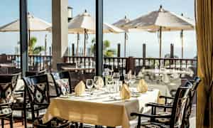 Et dækket bord i en restaurant med udsigt over en terrasse med parasoller og havet. (AI-genereret tekst)