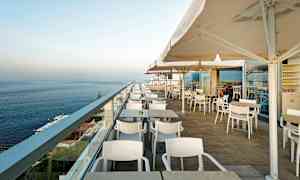 Billede af en udendørs terrasse med borde, stole og parasoller, med udsigt over havet. (AI-genereret tekst)