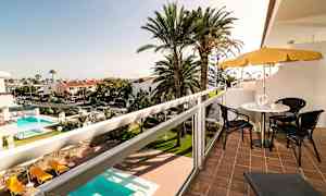 Udsigt fra en balkon med siddepladser og parasol, med en pool og palmer i baggrunden. (AI-genereret tekst)