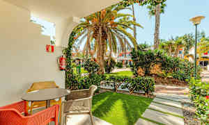 En terrasse med bord og farverige stole, indrammet af en bue, der fører ud til en have med palmer og bygninger i baggrunden. (AI-genereret tekst)