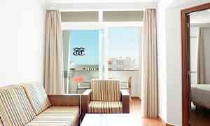 Et rum med en sofa og en lænestol beklædt med stribet stof, et sofabord og en balkon med udsigt til en bygning. (AI-genereret tekst)