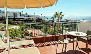 En terrasse med bord, stole og liggestole. I baggrunden ses palmer, bygninger og havet. (AI-genereret tekst)