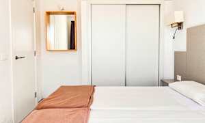 Et hotelværelse med hvidt interiør, en dobbeltseng med hvide lagner og brune sengetæpper samt et spejl på væggen. (AI-genereret tekst)