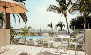 En altan med liggestole og et bord med stole, udsigt til en pool og palmer. (AI-genereret tekst)