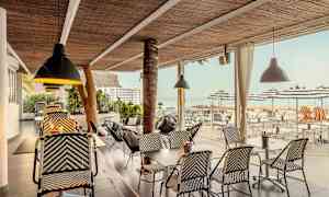 En udendørs servering med rottingmøbler og loftslamper, udsigt mod hav og parasoller. (AI-genereret tekst)