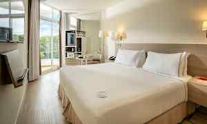 Hotelværelse med dobbeltseng og lys indretning, store vinduer og balkon med udsigt. (AI-genereret tekst)