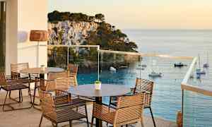 En terrasse med borde og stole, udsigt over havet og både ved en klippefyldt kyst. (AI-genereret tekst)