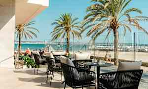 Udendørs serveringsområde med siddepladser og borde med udsigt til havet, palmer i baggrunden. (AI-genereret tekst)