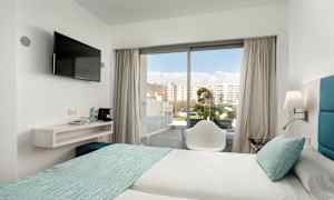 Et hotelværelse med to senge redt med lyse lagener og turkis sengetæppe. En stol står foran en glasdør, der fører ud til en altan. Et vægmonteret TV hænger ved siden af en hylde med vandflasker og kaffemaskine. (AI-genereret tekst)