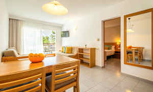 En lys stue med spisebord, stole, sofa og altan med udsigt til grønne områder. Et tilstødende soveværelse ses også. (AI-genereret tekst)