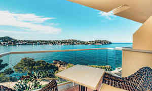 Udsigt fra en altan over en bugt med klart blåt vand, omgivet af kuperet terræn og træer. Et bord og stole ses i forgrunden på altanen. (AI-genereret tekst)