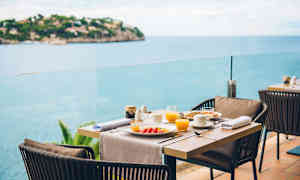 Dækket spisebord med tallerkener og glas på en terrasse med havudsigt. (AI-genereret tekst)