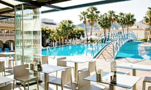 Et restaurantområde med udendørs servering ved en pool omgivet af palmer og bygninger. En blå gangbro strækker sig over poolen. (AI-genereret tekst)