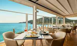 En restaurant med borde med mad og stole samt en panoramaudsigt over havet og en bjergkyst. (AI-genereret tekst)