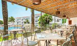 En udendørsrestaurant med borde og stole under et tag, med udsigt over en pool og hotelbygninger i baggrunden. (AI-genereret tekst)