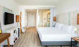 Et lyst hotelværelse med trådetailler og moderne møbler. Værelset har en stor seng, et tv og en åben planløsning. (AI-genereret tekst)