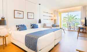 Et moderne hotelværelse med to senge og blå detaljer samt en altan med udsigt til palmer. (AI-genereret tekst)