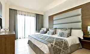 Et moderne soveværelse med en dobbeltseng, polstret sengegavl og udsmykkede tekstiler. Der er store glasdøre med lyse gardiner. (AI-genereret tekst)