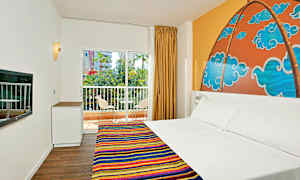 Et hotelværelse med en dobbeltseng og farverigt sengetæppe, et mønsteret vægmaleri bag sengen, et vindue med udsigt over altan og grønt. (AI-genereret tekst)