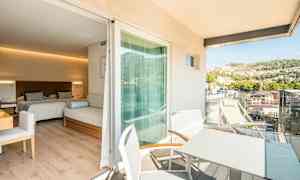 Et hotelværelse med to senge og en siddemøbel, med en tilhørende balkon hvor der er bord og stole samt udsigt over en frodig bakke. (AI-genereret tekst)