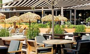 Et udendørsområde med borde, stole og liggestole under stråtag. (AI-genereret tekst)