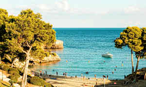 Strand med badende gæster og træer ved en klar blå hav, en båd er synlig på vandet. (AI-genereret tekst)