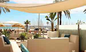 Udsigt over en afslappet siddegruppe med hvide møbler og blå puder, omgivet af tropiske planter, med en strand og en marina i baggrunden. (AI-genereret tekst)