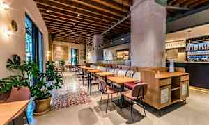 Et stilrent restaurantmiljø med trædetaljer i loftet, komfortable stole og borde samt dekorative planter. Et barområde ses i baggrunden. (AI-genereret tekst)