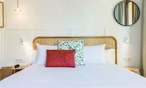 Et hotelværelse med en opredt dobbeltseng pyntet med hvide puder og en rød pyntepude foran en med blomsterdesign. Sengegavlen er lavet af flettet træ. Et rundt spejl og væglamper ses på en lys baggrund. (AI-genereret tekst)