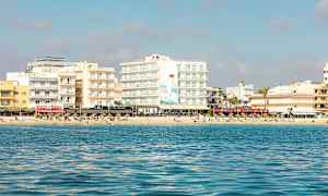 Udsigt fra havet mod en strand med flere bygninger og mennesker, der soler sig. (AI-genereret tekst)