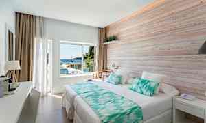 Et lyst hotelværelse med en stor seng, træpanel på væggen og et vindue med udsigt til havet. (AI-genereret tekst)