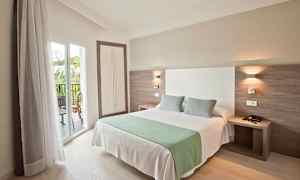 Et hotelværelse med en dobbeltseng, to puder og et grønt tæppe. Sengen er placeret tæt på en balkon med glasdøre. (AI-genereret tekst)