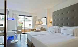 Hotelværelse med to senge, et vindue med udsigt til byen og et siddemiljø med bord og stol. (AI-genereret tekst)