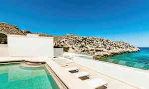 En udendørs pool ved et hus med udsigt over havet og klipper under en klar blå himmel. (AI-genereret tekst)