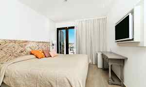 Et hotelværelse med en stor seng, dekorative puder, en vægmonteret TV og et skrivebord. Altandøre fører til en udsigt. (AI-genereret tekst)