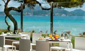 Billedet viser en terrasse med hvide borde og stole med udsigt over blåt hav og grønt område. (AI-genereret tekst)