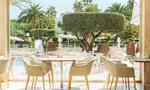 Restaurant med udsigt over udendørs pool og liggestole, omgivet af palmer og træer. (AI-genereret tekst)