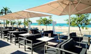 En udendørscafé med borde, stole og parasoller med udsigt over en sandstrand og havet. (AI-genereret tekst)