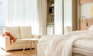 Et lyst hotelværelse med en sofa, et lille bord og en redt seng. I baggrunden er der et tekøkken med køleskab. (AI-genereret tekst)