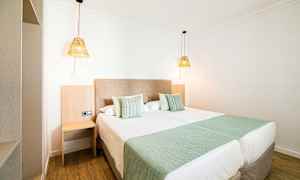 Et hotelværelse med to redte enkeltsenge, grønne sengetæpper og puder samt to hængende lamper. (AI-genereret tekst)