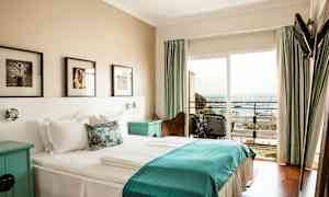 Et hotelværelse med en dobbeltseng og sengetøj i hvidt og blåt, vægkunst over sengen og en balkon med udsigt over havet. (AI-genereret tekst)