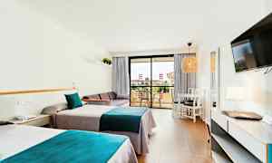 Et hotelværelse med to enkeltsenge med blå og grå sengetøj, en sofa, et bord med stole og en altan i baggrunden. (AI-genereret tekst)