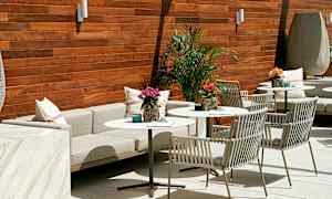 Et stilfuldt møblement på en terrasse med borde og stole, pyntet med planter og puder, mod en træpanelvæg. (AI-genereret tekst)